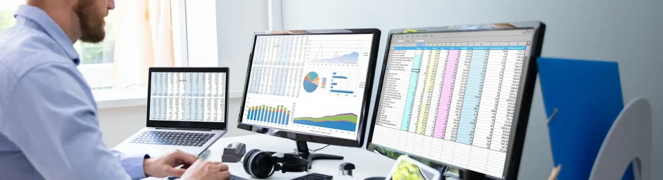 A man at a desk with three screens and lots of charts and data.